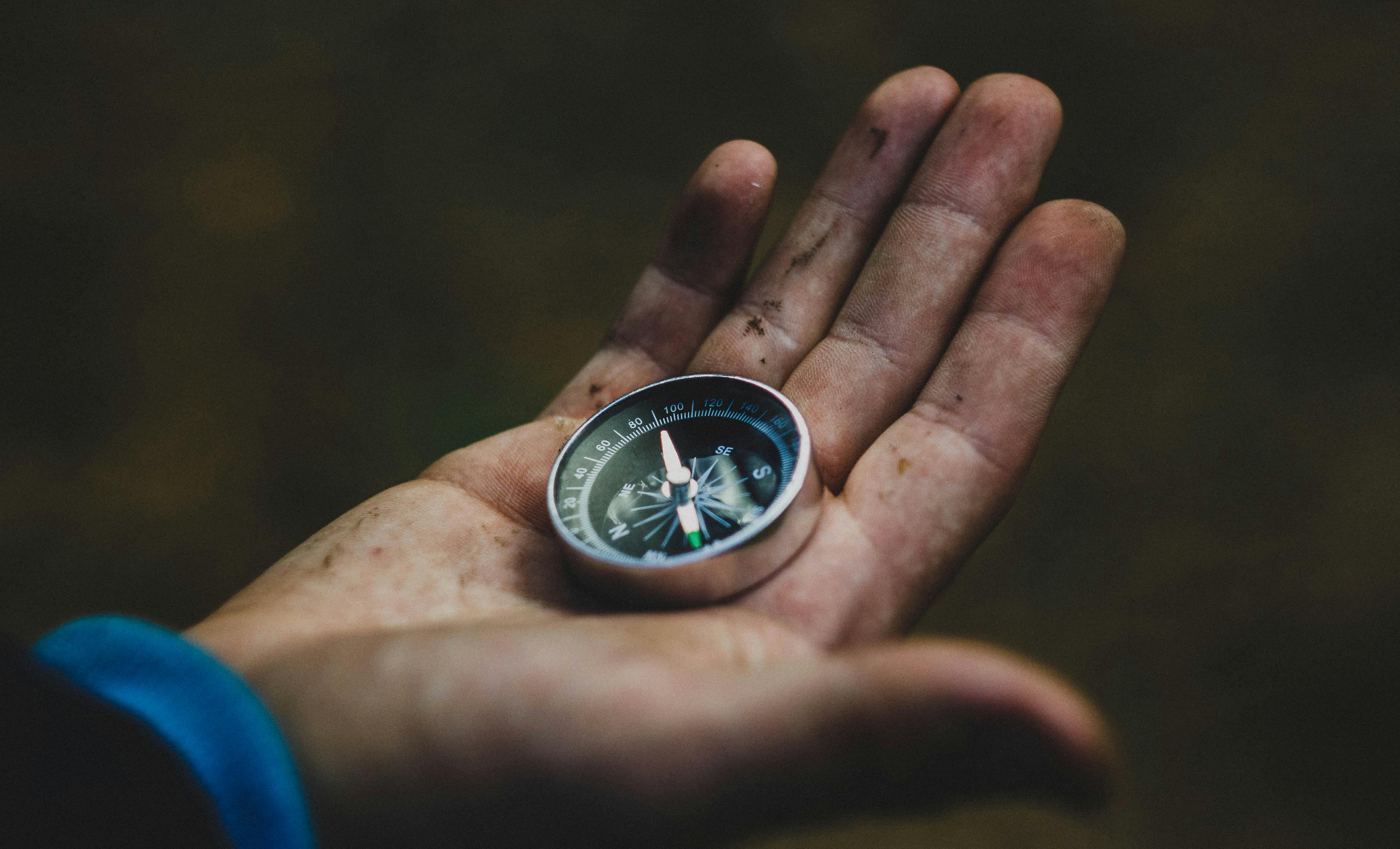 A photo of a compass, being held in a dirty hand by someone looking for the right direction to travel.